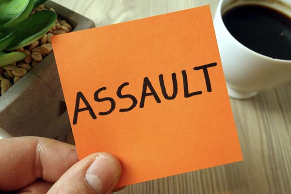 A hand holding an orange sticky note with the word ASSAULT written on it, with a coffee cup and plant in the background.