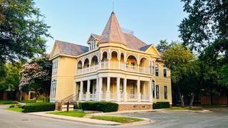 Large, historic yellow Victorian-style house with a prominent turret, arched wraparound porch, and two stories, located on a quiet, tree-lined street corner.