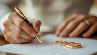 Close-up of a person signing a document with a gold pen, with two gold wedding rings placed on the paper in the foreground.