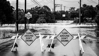 Flooded Texas road blocked by barricades and warning signs that read 'Turn Around Don't Drown,' with floodwaters covering the street and trees in the background.